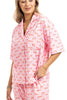 A woman beams in the Katydid Love Joy  Peace Preppy PJ Set—pink with red “love, joy, peace” print. The holiday pajamas include a short-sleeve collared button top with chest pocket and comfy pants with side pockets.