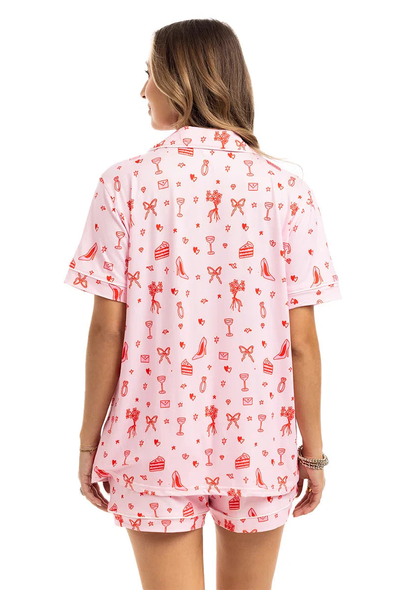 A woman with long hair is seen from behind wearing Katydids “Literally Just A Girl Icons” short pajama set in light pink, decorated with red bows, shoes, gifts, cupcakes, and drinks.