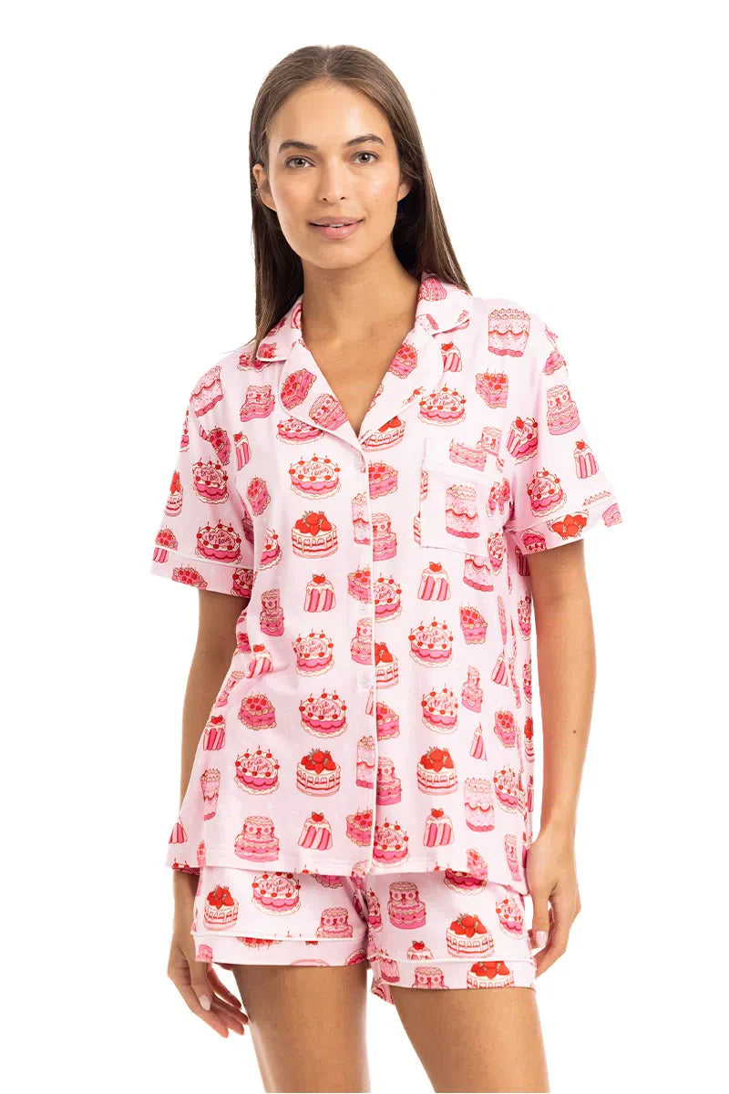 A woman wears the Katydid Girly Cakes PJ Shorts Set, featuring colorful cake and dessert prints, a short-sleeve button-up top, and matching shorts as she smiles at the camera.