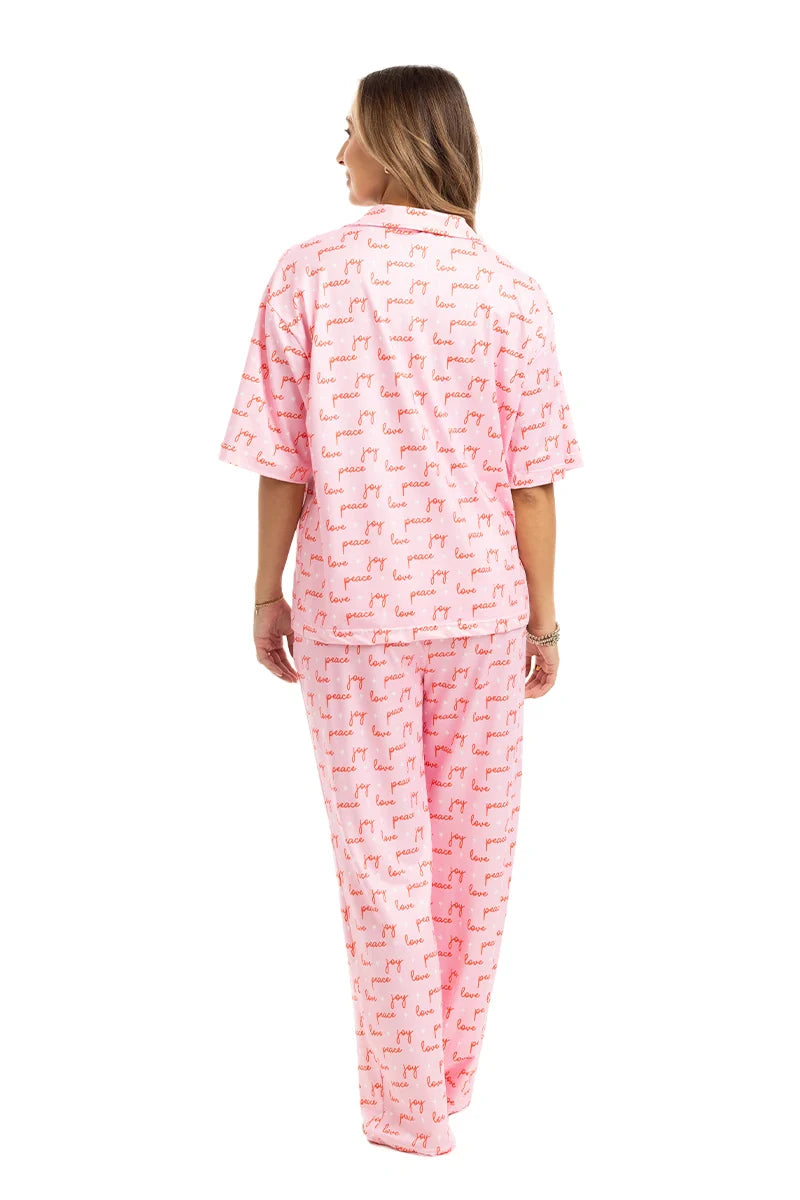 A woman with light brown hair stands facing away, wearing the Katydid Love Joy  Peace Preppy PJ Set. The festive set features a short-sleeve top and loose pink pants with red animal prints—a cozy Christmas pajama choice. White background.