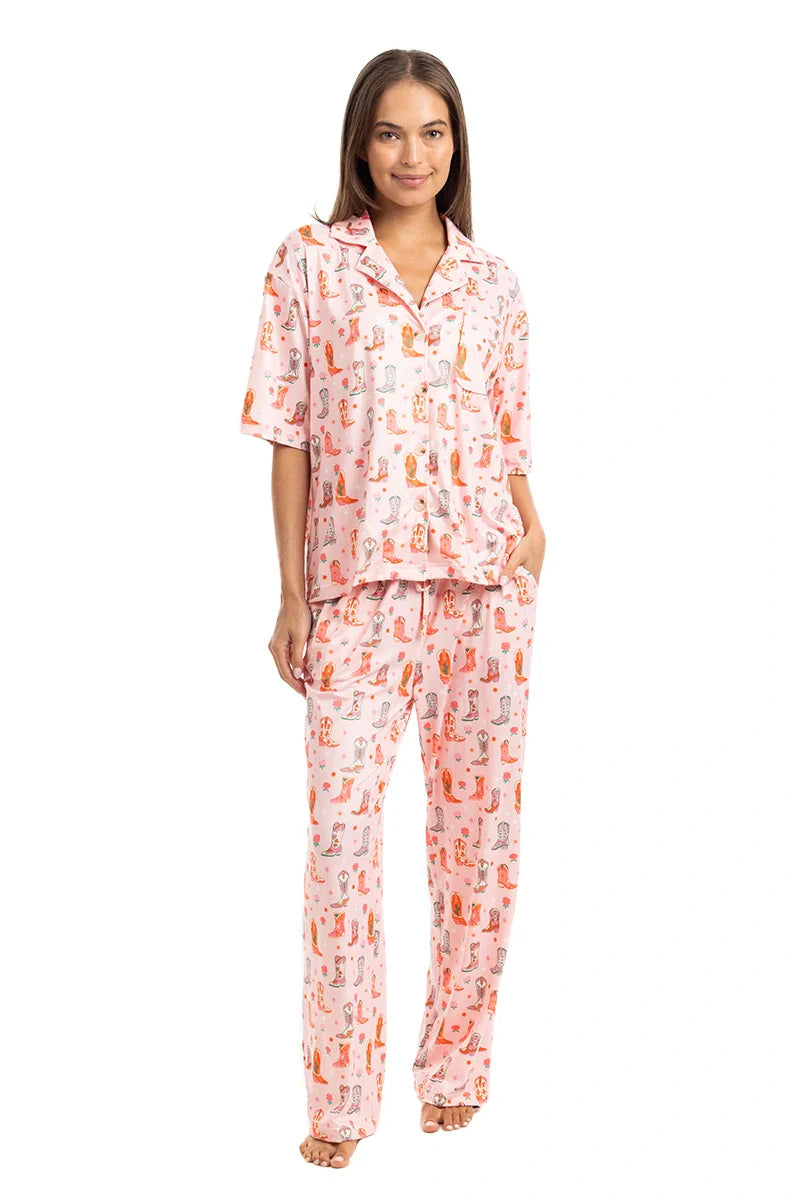 A woman smiles barefoot in the Katydid Womens Boots  Roses PJ Set—light pink with a colorful dog print. This set includes a button-up short-sleeve top and long pants, perfect playful cowgirl loungewear for relaxing at home.