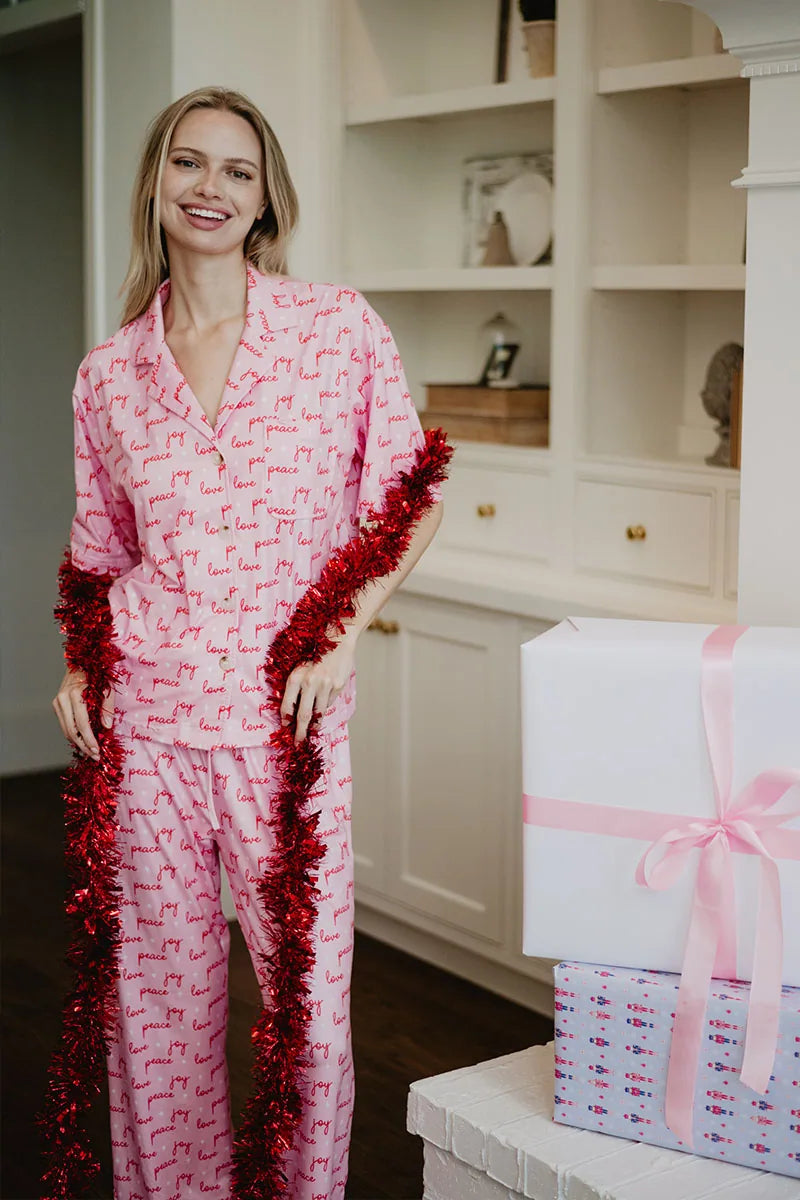 A woman in the Katydid Love Joy  Peace Preppy PJ Set smiles, holding red tinsel garland. Two presents with pink ribbons sit nearby among white shelves—a cozy Christmas scene perfect for festive holiday pajamas.