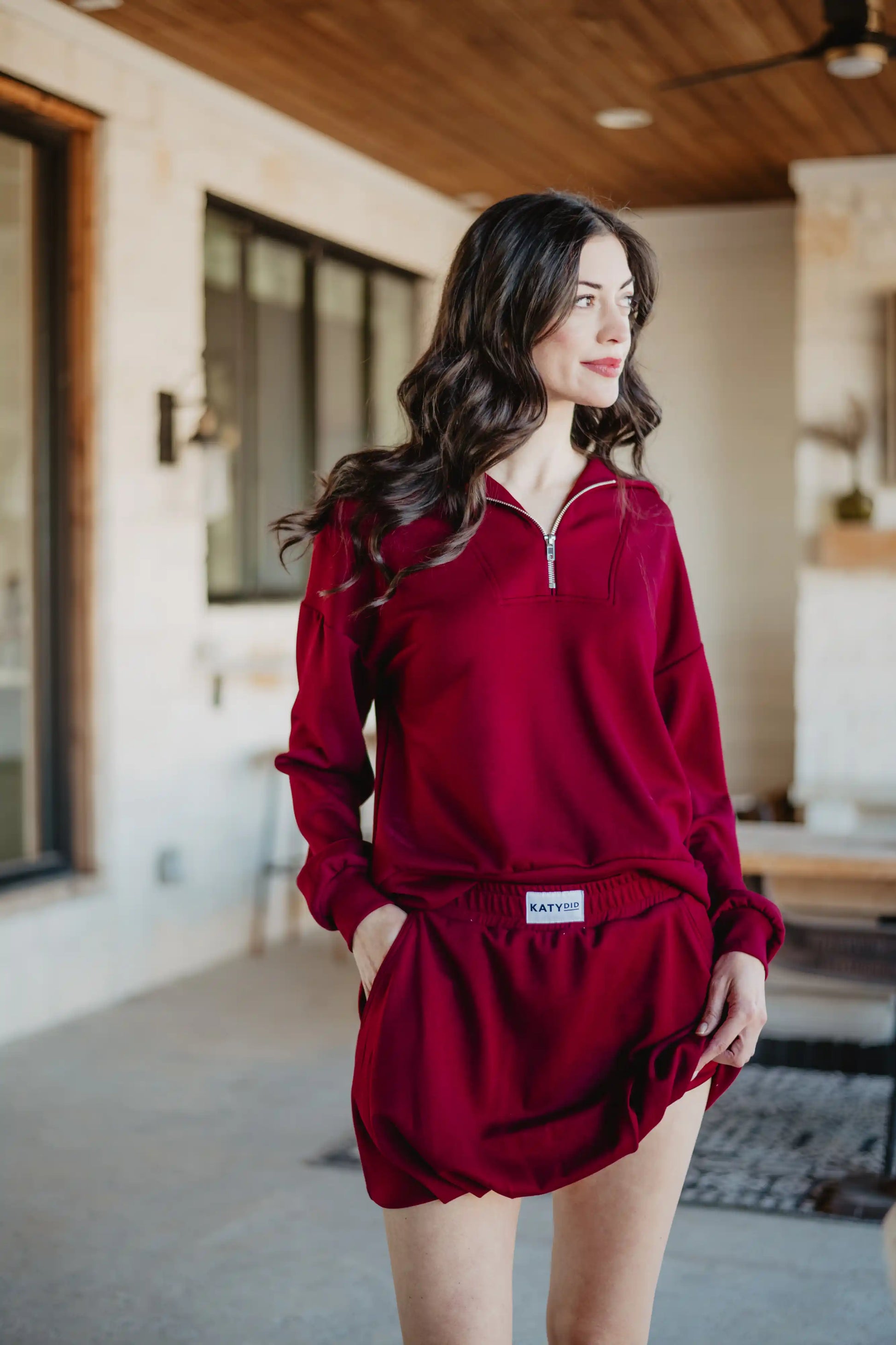 A woman with long, wavy brown hair wears the Katydid Wine Bubble Skirt Skort and a matching maroon zip-up sweatshirt, standing indoors by large windows with a relaxed expression.