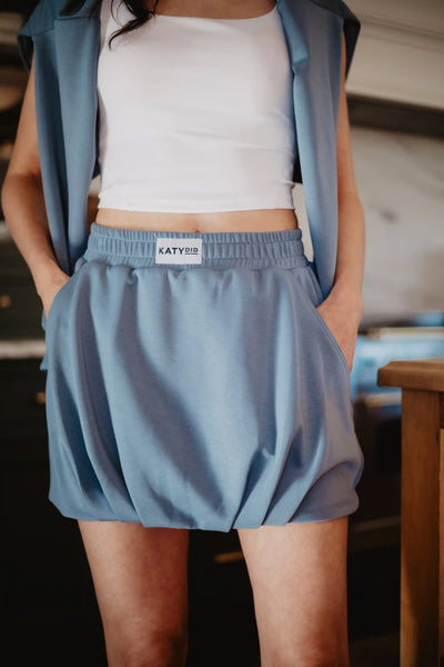 A person wearing a white crop top and a Katydid Steel Blue Mini Bubble Skirt with pockets; the skirt features a Katydid label on the waistband.