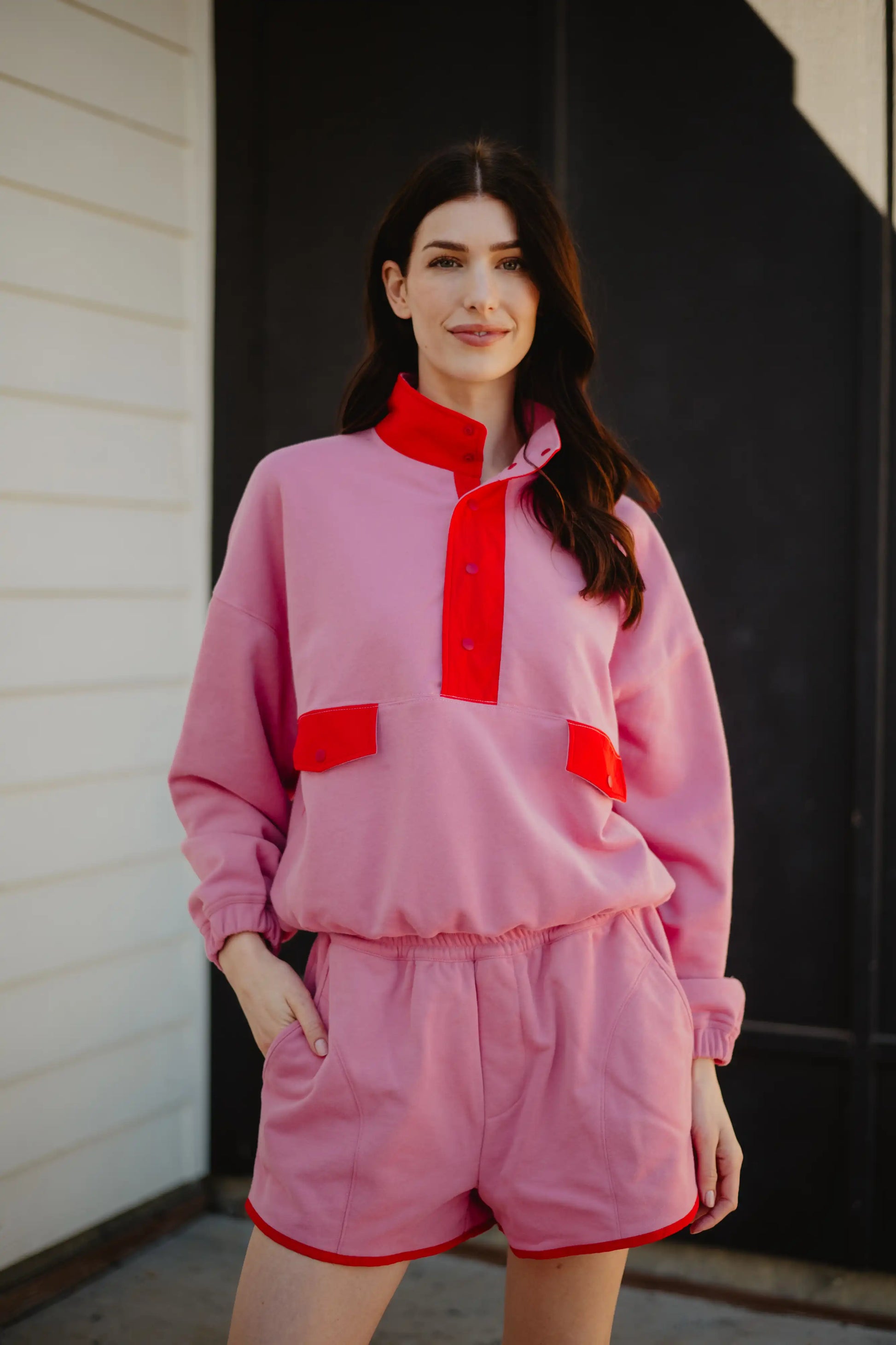 A woman stands outdoors in the Katydid Pink  Orange Quarter Snap 2 Piece Athletic Set, featuring a long-sleeve top with red accents and matching pink shorts, both crafted from ultra-soft performance fabric that complements her relaxed smile.