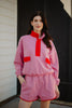 A woman stands outdoors in the Katydid Pink  Orange Quarter Snap 2 Piece Athletic Set, featuring a long-sleeve top with red accents and matching pink shorts, both crafted from ultra-soft performance fabric that complements her relaxed smile.