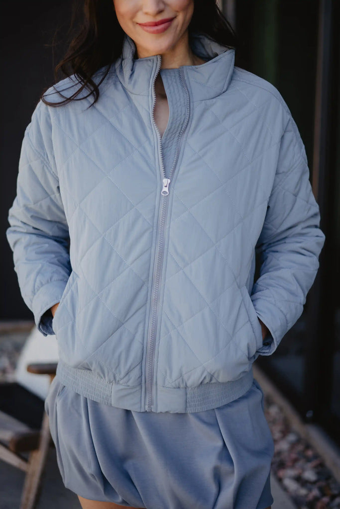 A woman stands outdoors, smiling slightly with hands in the pockets of a Katydid Steel Blue Sporty Diamond Quilted Womens Jacket, paired with a matching blue outfit.