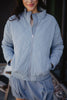 A woman stands outdoors, smiling slightly with hands in the pockets of a Katydid Steel Blue Sporty Diamond Quilted Womens Jacket, paired with a matching blue outfit.