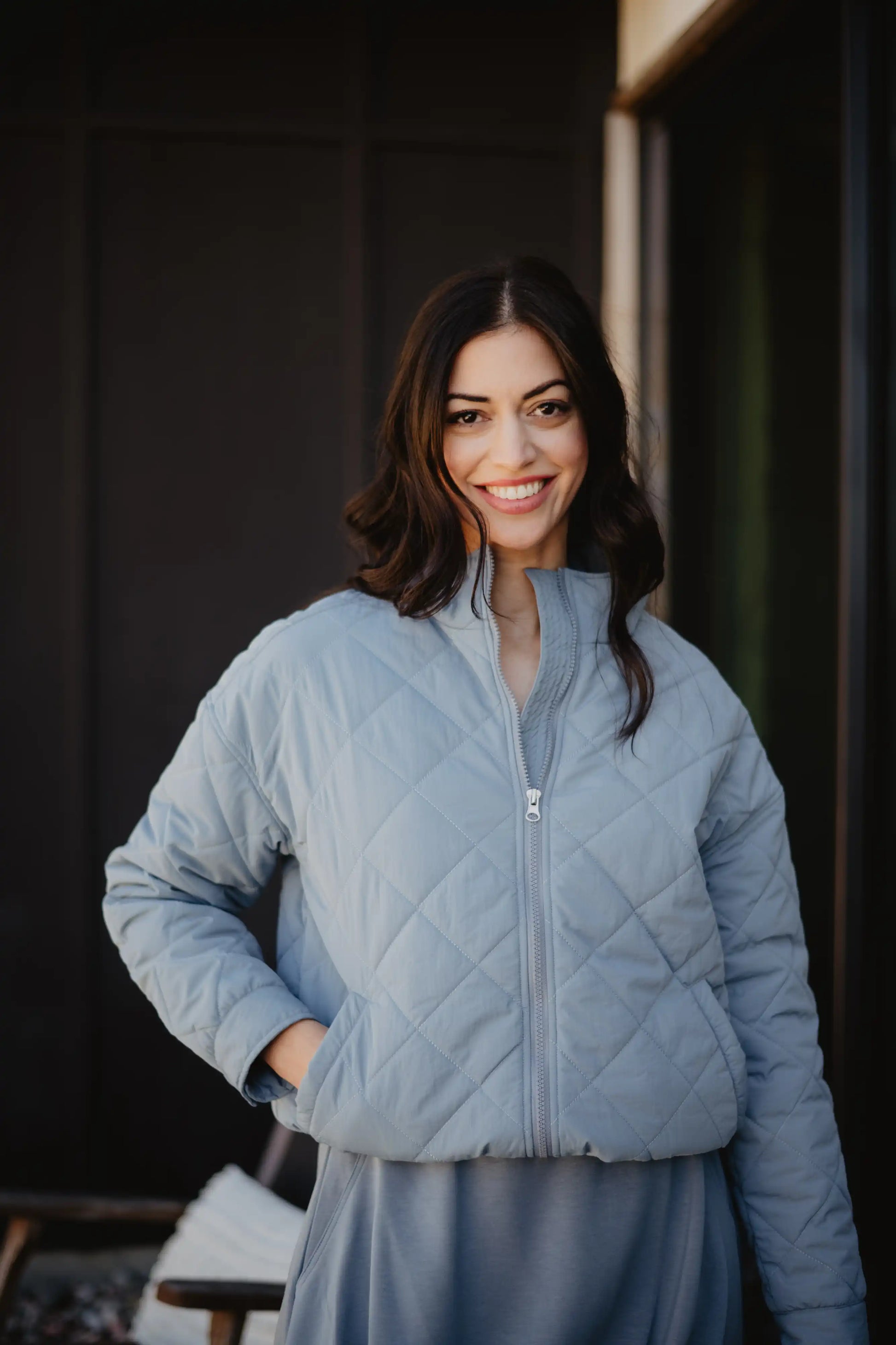 A woman with long dark hair smiles outdoors, wearing the Katydid Steel Blue Sporty Diamond Quilted Womens Jacket with one hand in her pocket. The background is dark and blurred.