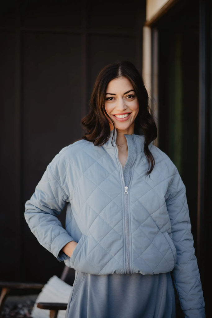 A woman with long dark hair smiles outdoors, wearing the Katydid Steel Blue Sporty Diamond Quilted Womens Jacket with one hand in her pocket. The background is dark and blurred.