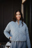 A woman with long dark hair smiles outdoors, wearing the Katydid Steel Blue Sporty Diamond Quilted Womens Jacket with one hand in her pocket. The background is dark and blurred.