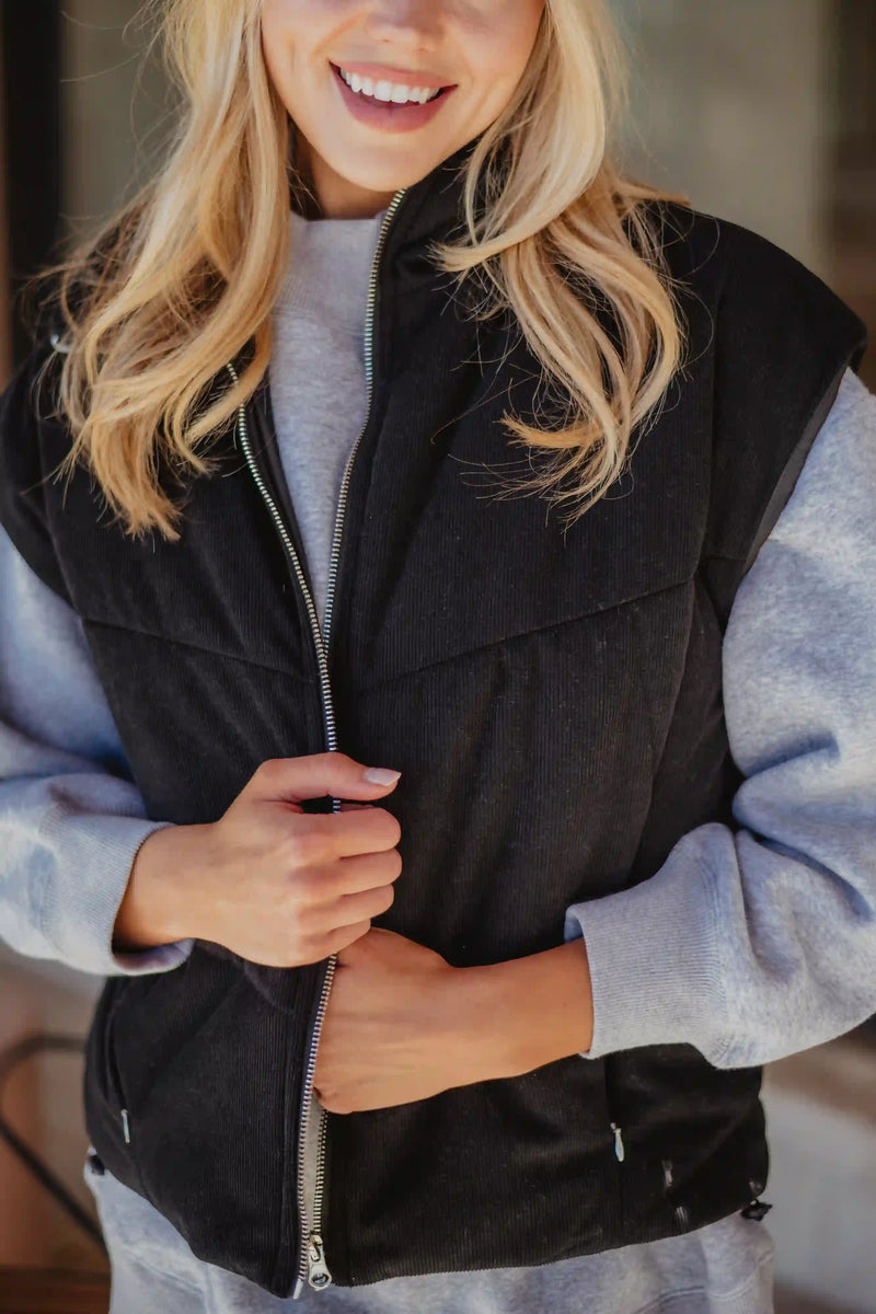 A smiling woman with long blonde hair models the Katydid Black Corduroy Trendy Puffer Vest over a gray sweatshirt, holding the vest near the zipper with both hands.