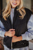 A smiling woman with long blonde hair models the Katydid Black Corduroy Trendy Puffer Vest over a gray sweatshirt, holding the vest near the zipper with both hands.