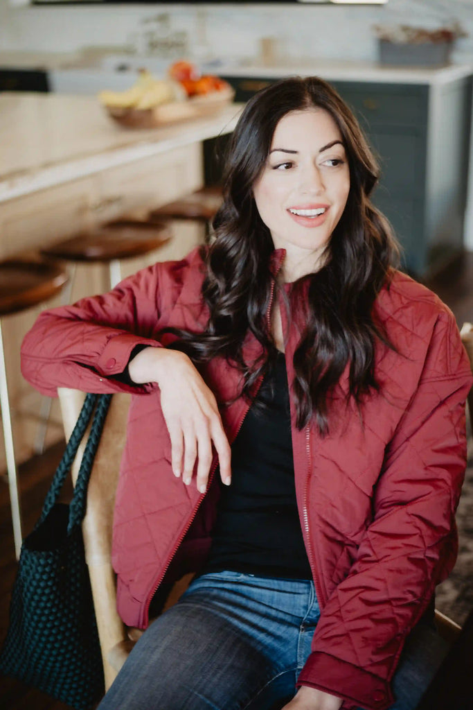 A woman with long dark hair sits indoors on a chair, smiling and looking to the side. She wears the Katydid Wine Diamond Quilted Jacket with jeans. Kitchen stools and a fruit-topped countertop are in the background.