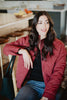 A woman with long dark hair sits indoors on a chair, smiling and looking to the side. She wears the Katydid Wine Diamond Quilted Jacket with jeans. Kitchen stools and a fruit-topped countertop are in the background.