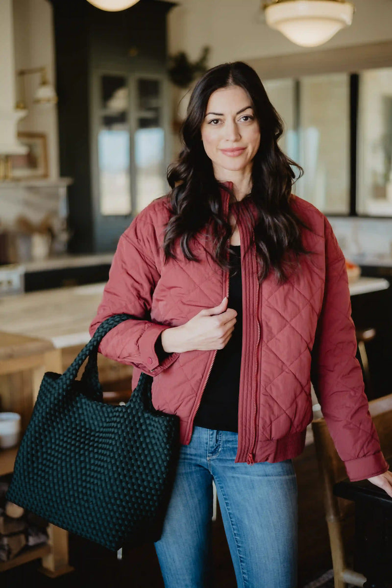 A woman with long dark hair wears the Katydid Wine Diamond Quilted Jacket and blue jeans indoors, holding a textured black tote bag and smiling softly at the camera.