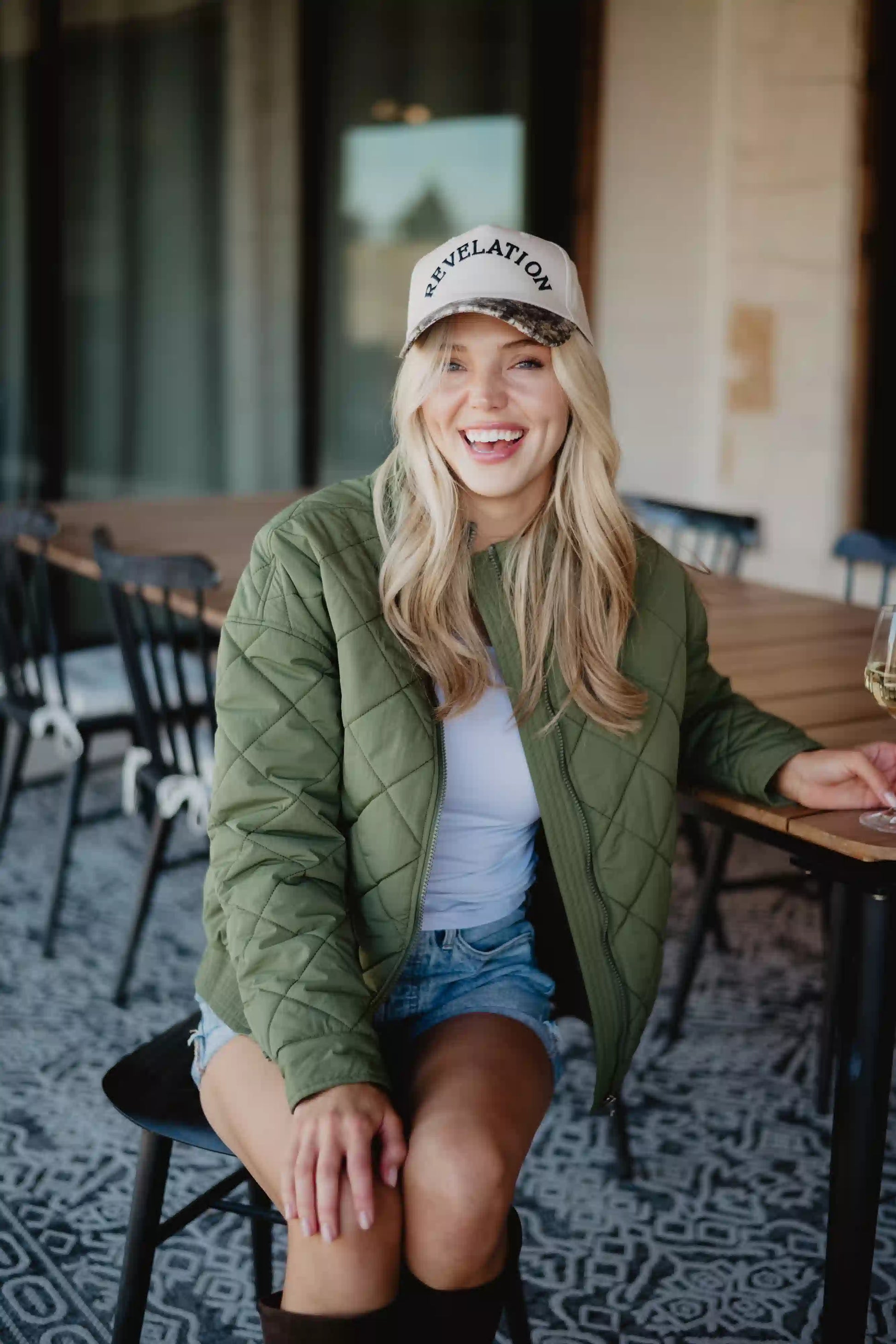 A smiling woman with long blonde hair wears the Katydid Olive Sporty Diamond Quilted Jacket as she sits outdoors on a black chair by a table, paired with a white shirt, denim shorts, and a REVELATION cap.