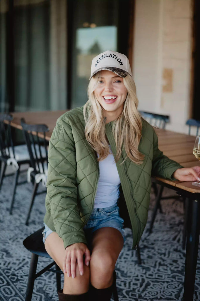 A smiling woman with long blonde hair wears the Katydid Olive Sporty Diamond Quilted Jacket as she sits outdoors on a black chair by a table, paired with a white shirt, denim shorts, and a REVELATION cap.