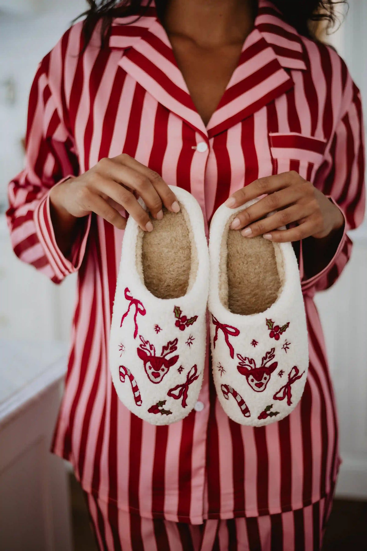 Wearing red and pink striped pajamas, someone holds Katydid’s Bow Reindeer Winter Slippers—fuzzy white sherpa slippers decorated with red reindeer, candy canes, bows, and stars.