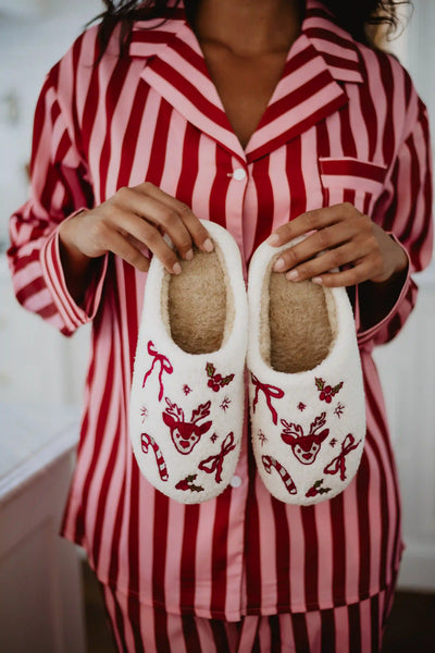 Wearing red and pink striped pajamas, someone holds Katydid’s Bow Reindeer Winter Slippers—fuzzy white sherpa slippers decorated with red reindeer, candy canes, bows, and stars.