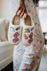 Wearing pastel-patterned pajamas, someone holds Katydid’s Mahjong Pieces Cozy Slippers, featuring colorful embroidered fans and flowers. A softly blurred background hints at a warm, comfy indoor setting.
