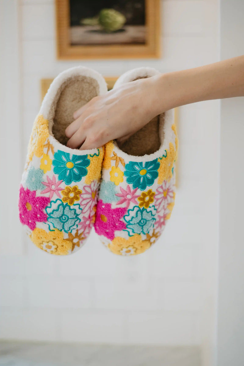 A hand holds the Katydid Spring Flowers House Slippers for Women, featuring colorful blue, pink, and yellow floral patterns, against a light indoor background.