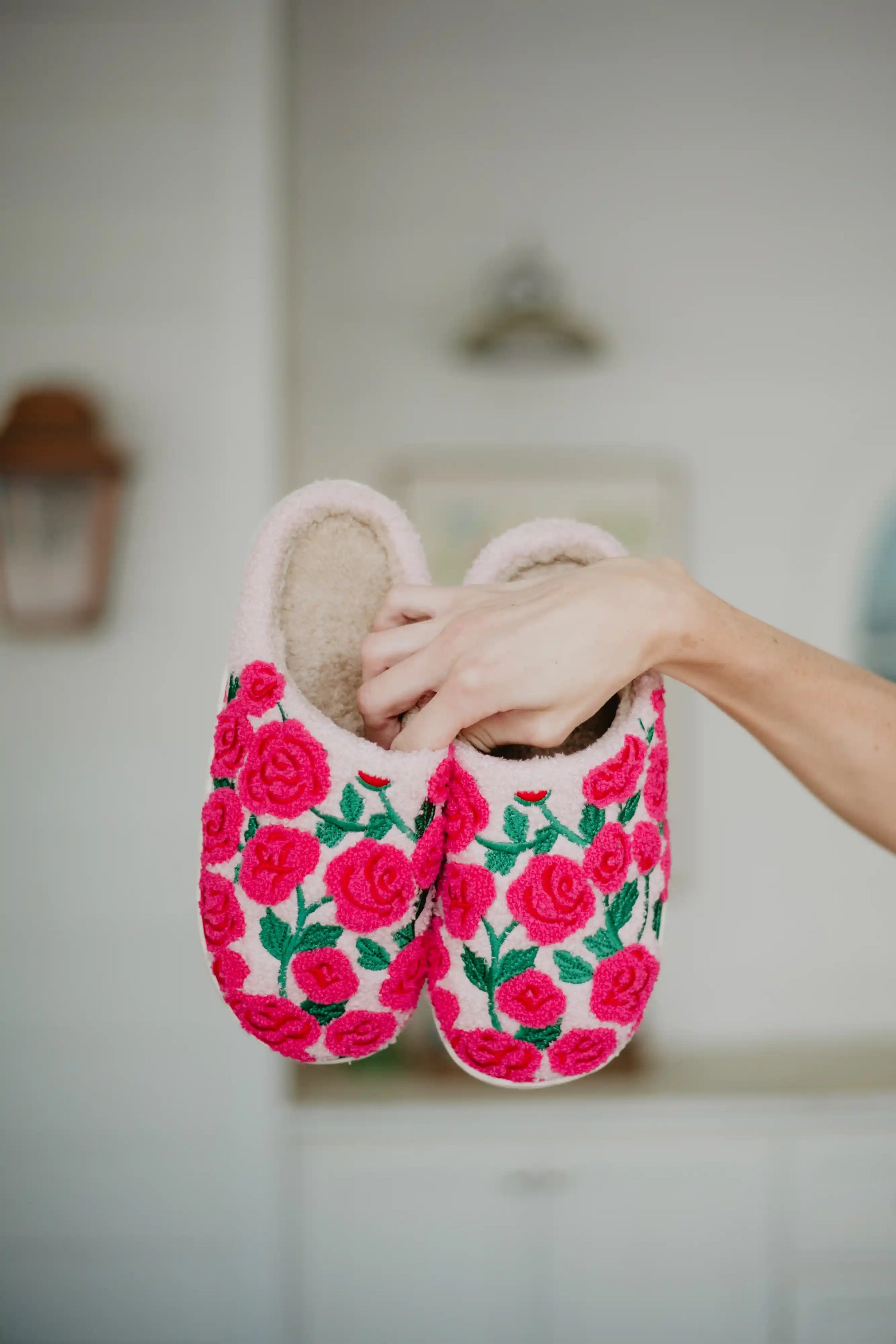 A hand displays Katydid’s Whimsical Roses Fuzzy Slippers for women, featuring bright pink embroidered roses and green leaves, against a softly blurred indoor background.
