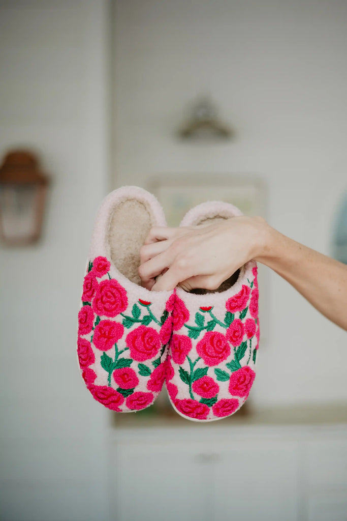 A hand displays Katydid’s Whimsical Roses Fuzzy Slippers for women, featuring bright pink embroidered roses and green leaves, against a softly blurred indoor background.