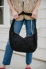 A person in blue jeans and a beige quilted vest holds the Katydid Black Spacious Suede Tote Bag, a stylish womens handbag for fall, against a light-colored wall.
