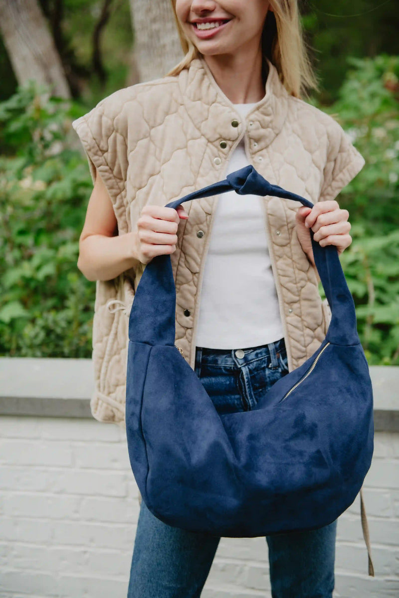 A person in a beige quilted vest, white top, and jeans holds the Katydid Blue Suede Evening Tote by its handle while standing outdoors with greenery behind them.