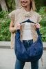 A person in a beige quilted vest, white top, and jeans holds the Katydid Blue Suede Evening Tote by its handle while standing outdoors with greenery behind them.