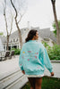 A woman smiles outside a large house, looking to the side while wearing a light blue Katydid Make Heaven Crowded Womens Mockneck Top featuring colorful designs and lettering on the back.