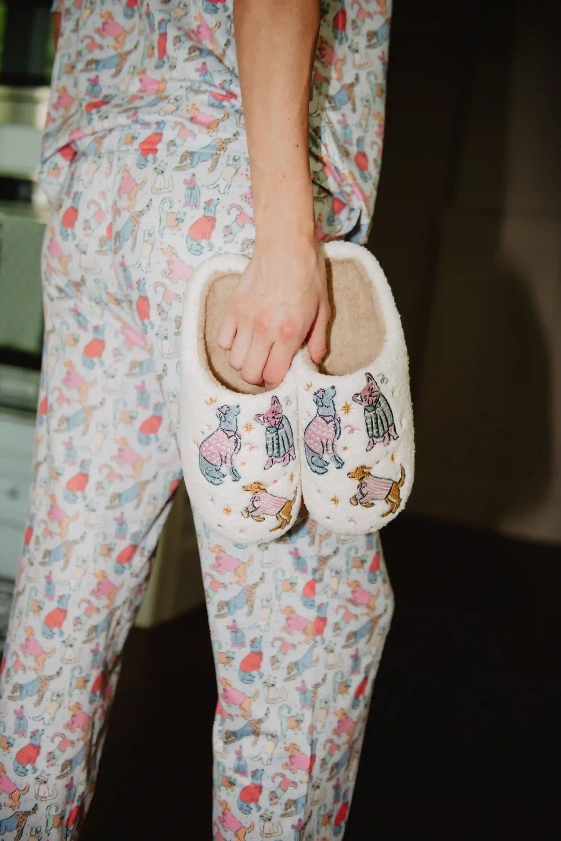 Wearing pastel animal-print pajamas, a woman holds Katydid’s Posh Pets Women Slippers—fuzzy sherpa footwear featuring cute, colorfully dressed animals—for a cozy, relaxed vibe indoors.