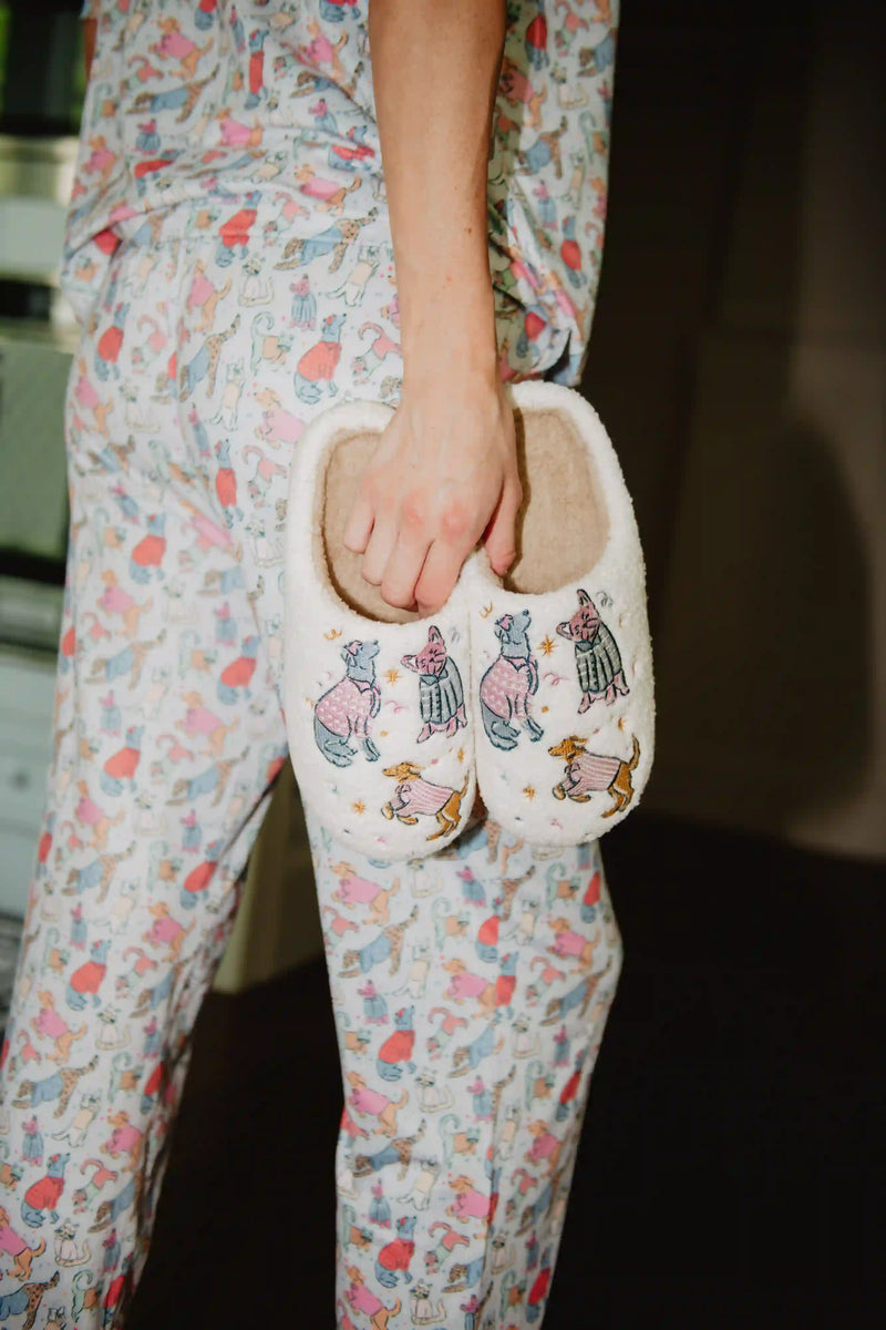 Wearing pastel animal-print pajamas, a woman holds Katydid’s Posh Pets Women Slippers—fuzzy sherpa footwear featuring cute, colorfully dressed animals—for a cozy, relaxed vibe indoors.