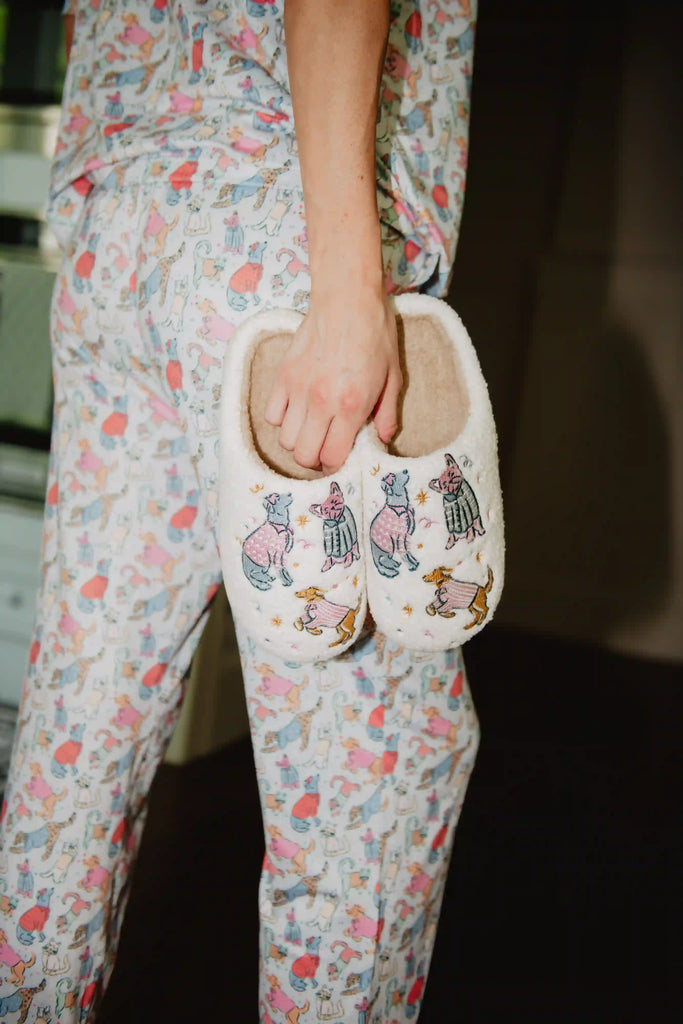 Wearing pastel animal-print pajamas, a woman holds Katydid’s Posh Pets Women Slippers—fuzzy sherpa footwear featuring cute, colorfully dressed animals—for a cozy, relaxed vibe indoors.