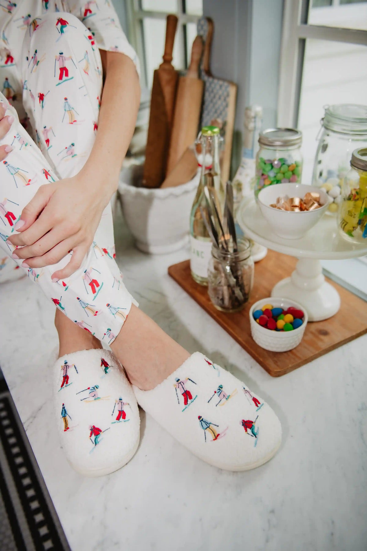 Wearing Katydid Apres Ski Winter Slippers and colorful pajama pants, a person sits on a kitchen counter near jars of candy, glass bottles, a bowl of colorful sweets, and wooden utensils, with their hands resting on their knee.