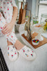 Wearing Katydid Apres Ski Winter Slippers and colorful pajama pants, a person sits on a kitchen counter near jars of candy, glass bottles, a bowl of colorful sweets, and wooden utensils, with their hands resting on their knee.