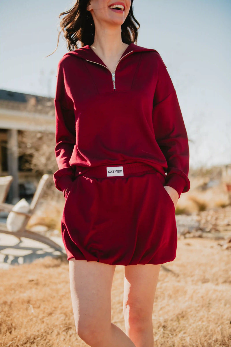 A woman smiles outdoors, hands in her pockets, wearing a matching maroon zip-up sweatshirt and the Katydid Wine Bubble Skirt Skort with visible branding. The setting is sunny with a slightly blurred background.