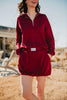 A woman smiles outdoors, hands in her pockets, wearing a matching maroon zip-up sweatshirt and the Katydid Wine Bubble Skirt Skort with visible branding. The setting is sunny with a slightly blurred background.