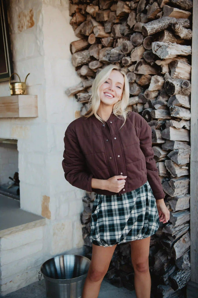 A smiling woman with blonde hair wears the Katydid Chocolate Brown Cropped Quilted Bomber Jacket, paired with a green plaid skirt, in front of a woodpile and stone wall.