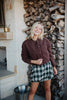 A smiling woman with blonde hair wears the Katydid Chocolate Brown Cropped Quilted Bomber Jacket, paired with a green plaid skirt, in front of a woodpile and stone wall.