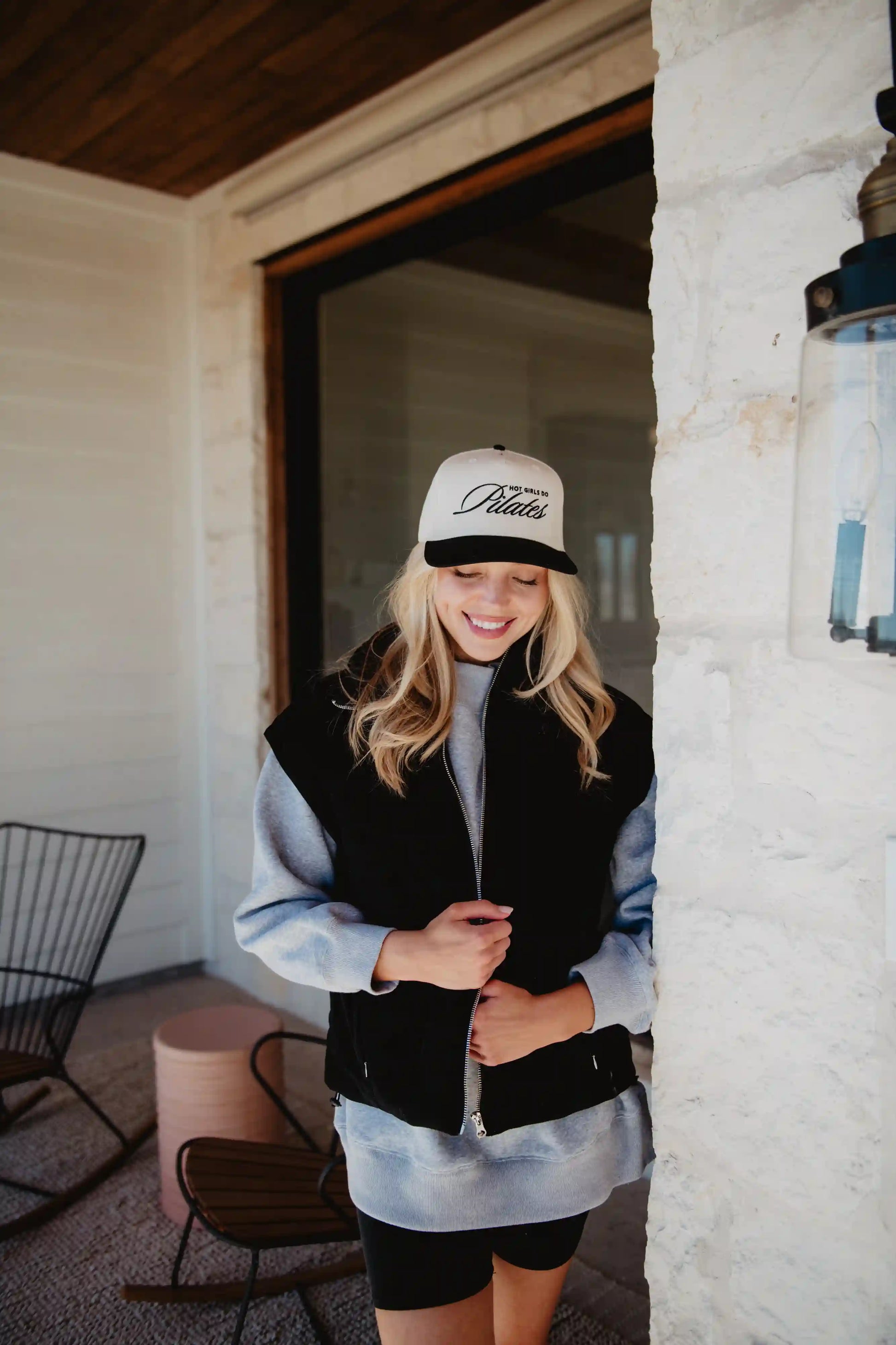 A woman with long blonde hair smiles and looks down near a white brick wall, wearing a Katydid Black Corduroy Trendy Puffer Vest, a light hoodie, and a white baseball cap. Patio furniture is visible in the background.