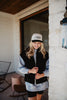 A woman with long blonde hair smiles and looks down near a white brick wall, wearing a Katydid Black Corduroy Trendy Puffer Vest, a light hoodie, and a white baseball cap. Patio furniture is visible in the background.