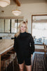 A woman with blonde hair smiles in a modern kitchen, wearing the Katydid Black Mockneck Sweatshirt. The space features a marble countertop, wooden stools, and large windows filling the room with natural light.