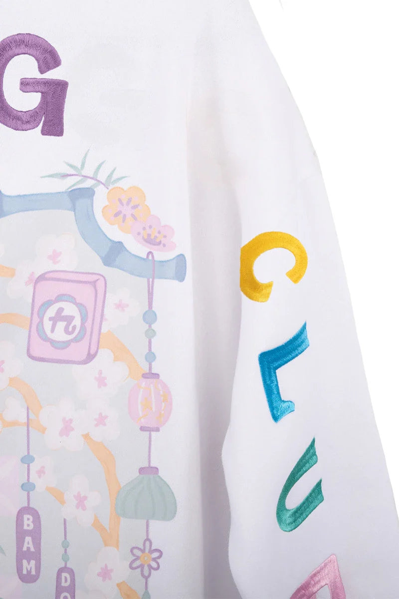A close-up of the Katydid Mahjong Club Mockneck Pullover, a white sweatshirt with colorful G, C, E, and A letters on the sleeve and pastel floral and decorative patterns on the fabric.