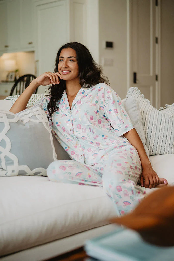 A woman wearing the Katydid Mahjong Pieces Pajama Pants Set smiles comfortably on a white couch, resting her arm on a cushion in a bright, cozy living room.
