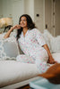 A woman wearing the Katydid Mahjong Pieces Pajama Pants Set smiles comfortably on a white couch, resting her arm on a cushion in a bright, cozy living room.