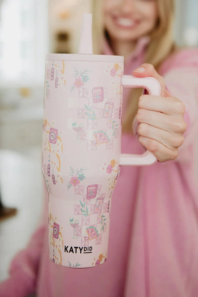 A smiling woman in a pink top holds the Katydid Mahjong Pieces Hydration Tumbler, featuring floral and envelope motifs, in a welcoming indoor setting.