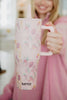 A smiling woman in a pink top holds the Katydid Mahjong Pieces Hydration Tumbler, featuring floral and envelope motifs, in a welcoming indoor setting.