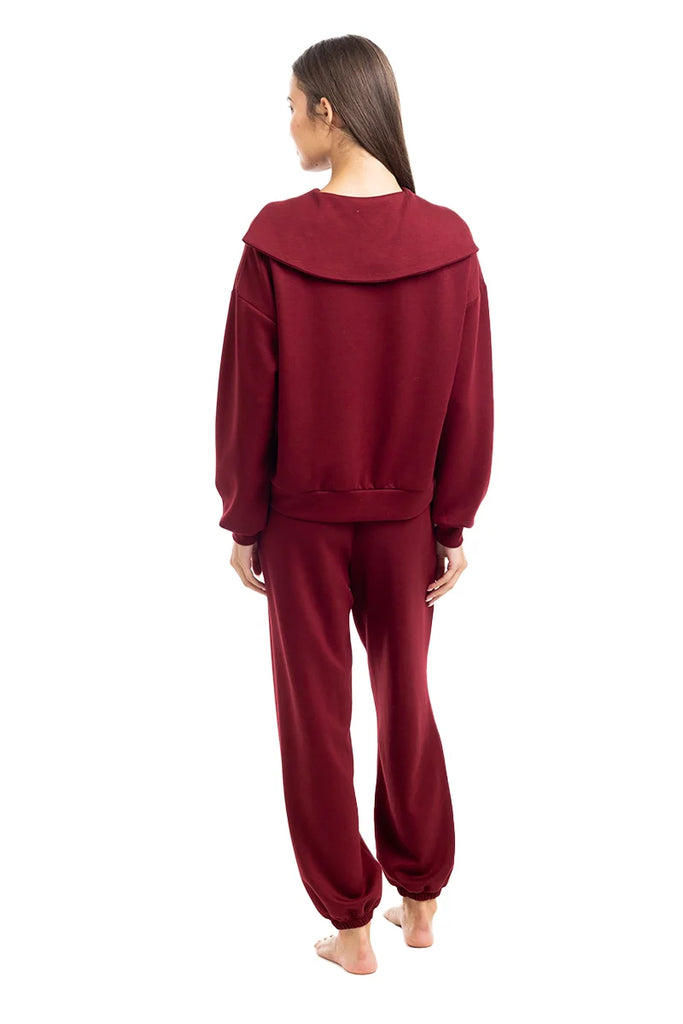 A woman with long brown hair, barefoot and facing away, showcases the Katydid Wine Pants Two-Piece Set with a cozy maroon wide-collar sweatshirt and matching pants against a plain white background.